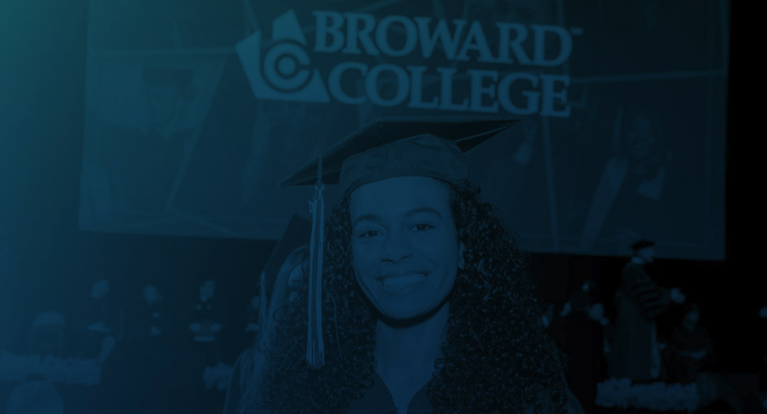 Black female graduate smiling, with the Broward College logo on a large screen in the background.